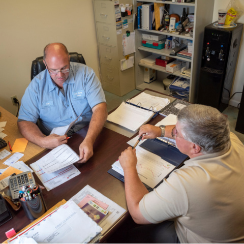 A Purina nutritionist consults with a dairy farmer in an office, both men looking at papers and charts. 