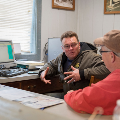 Two male dairy farmers sit in a farm office talking with a computer and papers on the desk.